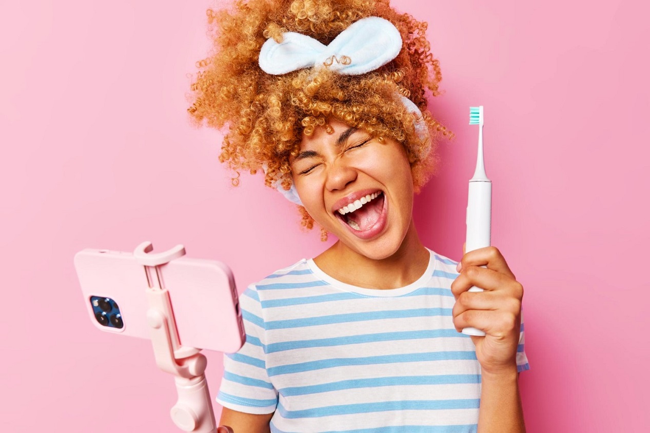 Jeune femme métissée se brossant les dents avec une brosse à dents électrique en regardant son smartphone